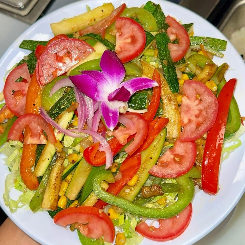 Colorful vegetable salad with tomatoes, zucchini, peppers, corn, onions, lettuce, and a purple orchid garnish on a white plate.