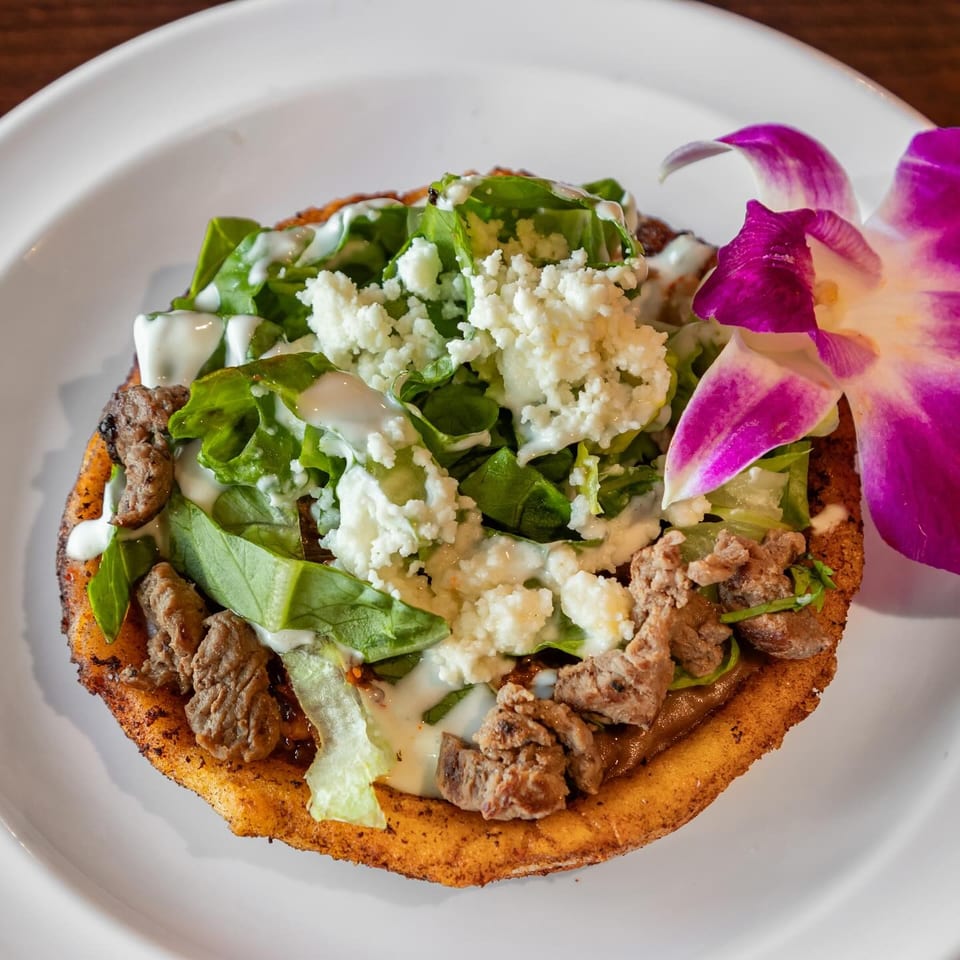 Crispy flatbread topped with grilled meat, leafy greens, crumbled cheese, creamy dressing, and a purple orchid garnish.