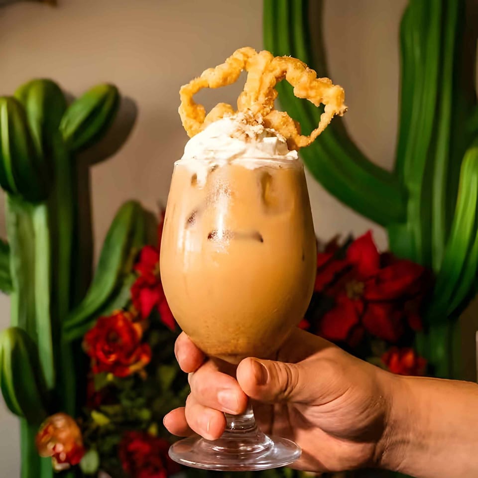Creamy orange dessert cocktail topped with whipped cream and fried garnish, held in a stemmed glass against tropical foliage.