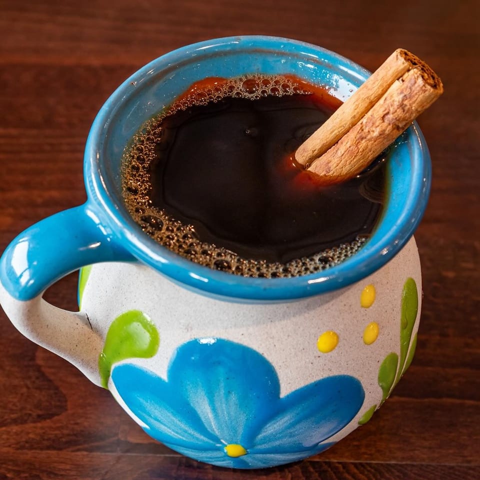 Dark coffee served in a colorful hand-painted ceramic mug, garnished with two cinnamon sticks on a wooden table.