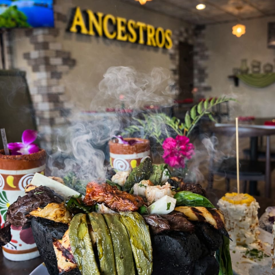 Steaming grilled Mexican-style platter served in a black volcanic stone bowl, garnished with vegetables and flowers at Ancestros restaurant.