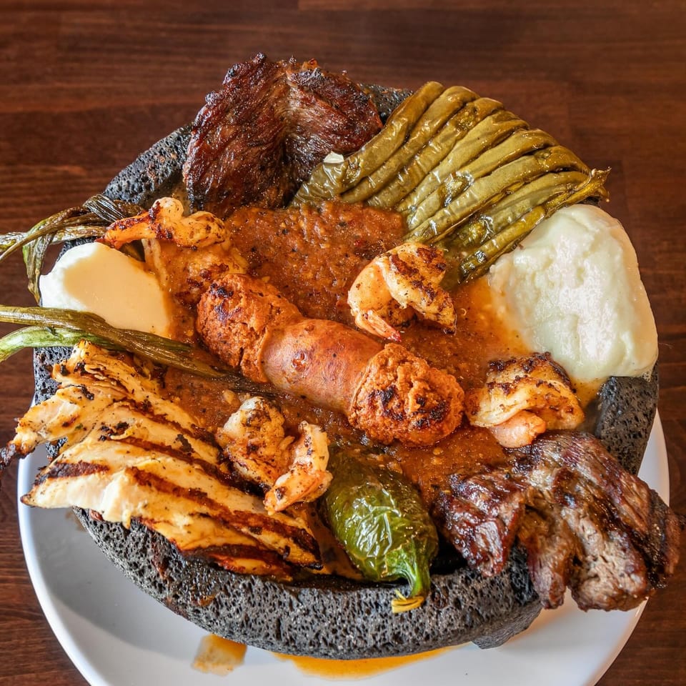 Loaded platter of grilled meats, shrimp, sausage, chicken, vegetables, and creamy sides served on a black stone plate.