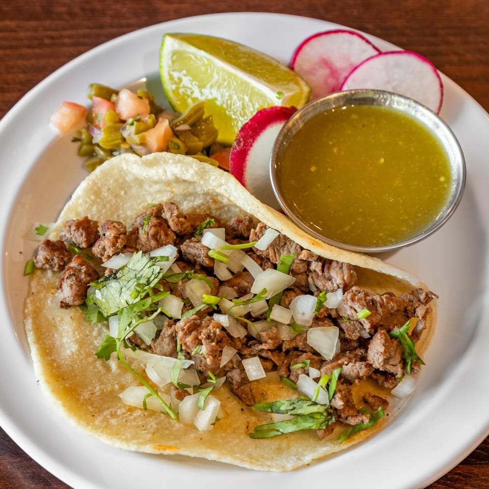 Mexican steak tacos topped with onions and cilantro, served with green salsa, lime, radishes, and pickled vegetables.