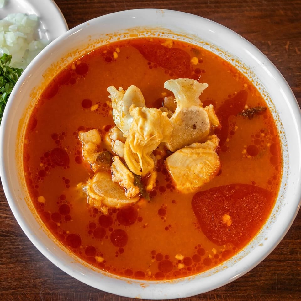 Bowl of spicy red broth with chicken, vegetables, and a large corn tortilla chip, served with chopped onion and cilantro.